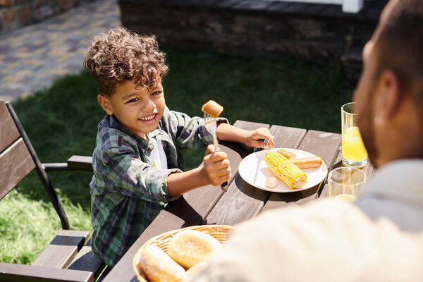 joyful curly african american boy eating sausages and grilled corn while looking at father outdoors