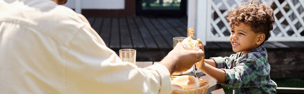happy african american father passing fresh bun to curly smiling son while having family bbq, banner