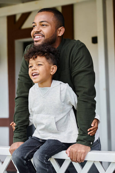 joyful african american boy sitting on porch and embracing his father in braces on backyard of house