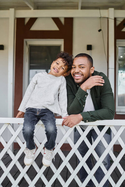 positive african american father and son smiling and sitting on porch of house, family portrait