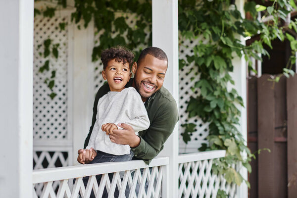 happy african american man in braces laughing and hugging curly son while sitting on porch
