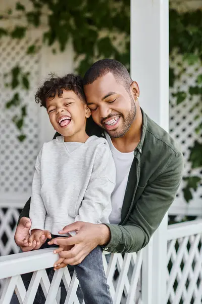 Beyaz çitli verandada oturmuş kıvırcık oğluna sarılan, diş teli takan mutlu Afro-Amerikalı adam.