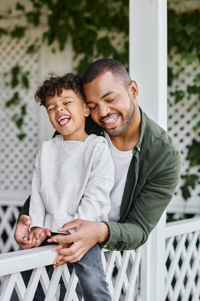 happy african american man in braces hugging curly son while sitting on porch with white fence