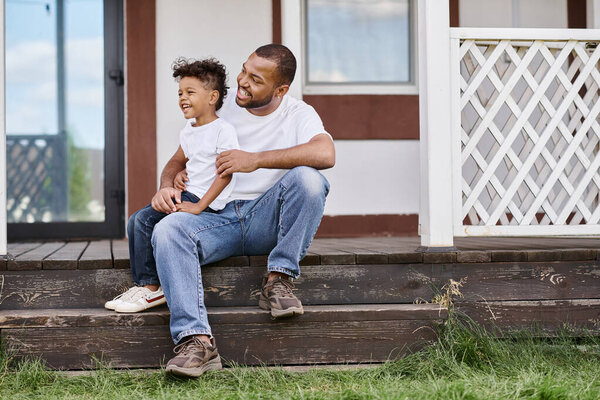 cheerful african american man in braces hugging curly son while sitting on porch of modern house