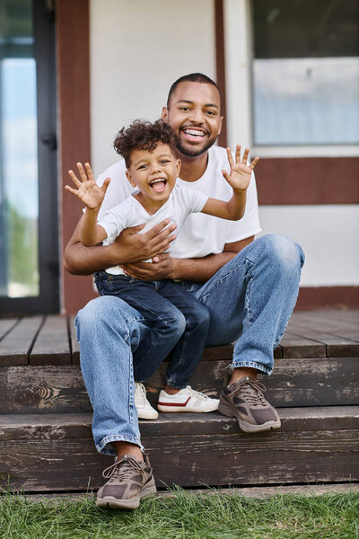 cheerful african american man in braces hugging excited son while sitting on porch of modern house