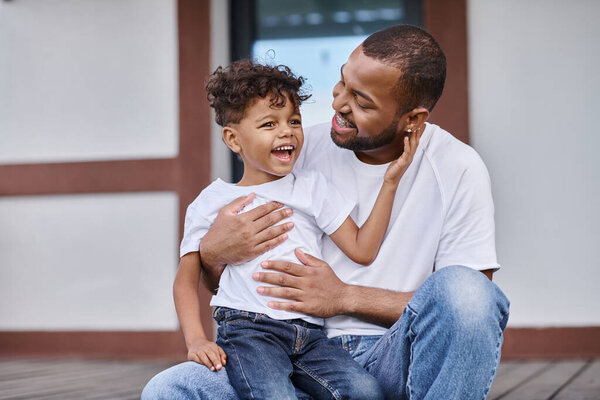 jolly african american boy hugging happy father in braces while sitting on porch of modern house