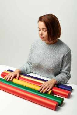 Kısa saçlı genç bir kadın masasında gri arkaplanlı renkli hediye kağıdına bakıyor.