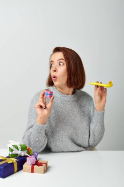 shocked woman in sweater holding tiny Christmas gift and yellow party horn on grey backdrop clipart