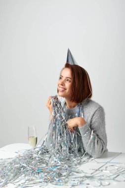 satisfied woman in party cap and tinsel smiling near champagne glass while celebrating New year clipart