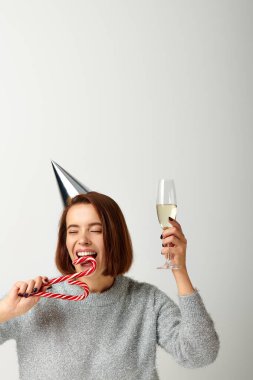 Parti şapkalı genç kadın elinde şampanya bardağı ve gri Noel 'de şeker kamışı ısırıyor.