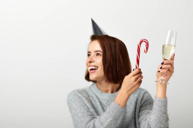 joyful woman in party cap holding champagne glass and candy cane on grey, Merry Christmas concept clipart