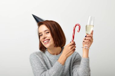 positive woman in party cap holding champagne glass and candy cane on grey, Merry Christmas clipart