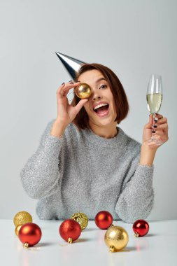 excited woman in party cap holding champagne glass and bauble on grey, Merry Christmas concept clipart