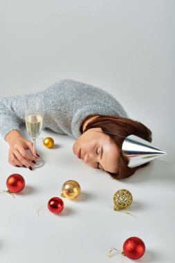 Parti şapkalı kadın şampanya bardağının yanında uyuyor ve Noel mücevherleri gri arka planda.