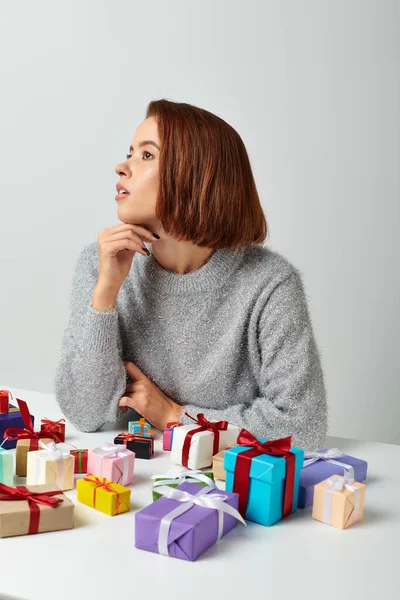 stock image dreamy woman in winter sweater fantasizing among bunch of Christmas gifts on table, grey backdrop