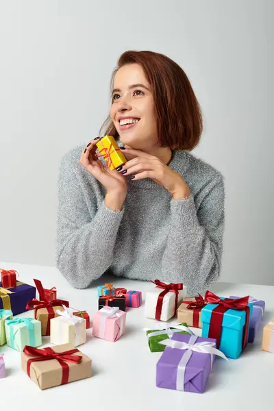 stock image cheerful woman in sweater fantasizing and holding Christmas gift near bunch of presents on table