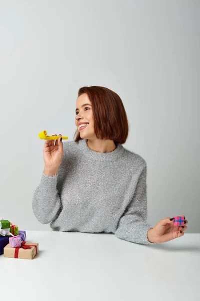 stock image cheerful woman in sweater fantasizing and holding tiny Christmas gift and yellow party horn on grey