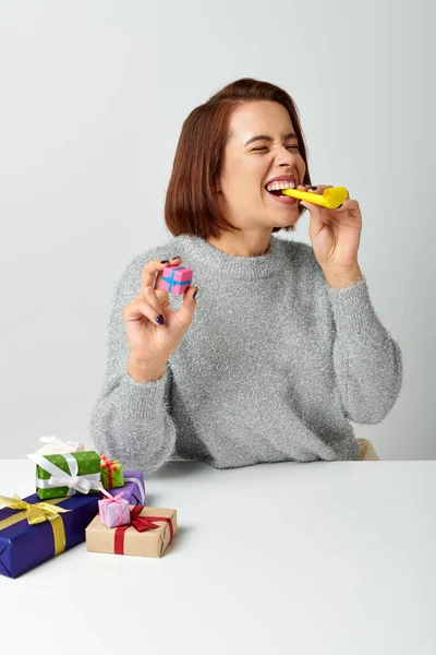 happy woman in sweater holding tiny Christmas gift and blowing yellow party horn on grey backdrop