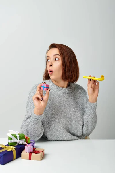 shocked woman in sweater holding tiny Christmas gift and yellow party horn on grey backdrop