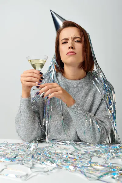 stock image serious woman in party cap with tinsel on head holding glass of champagne on grey, New Year concept