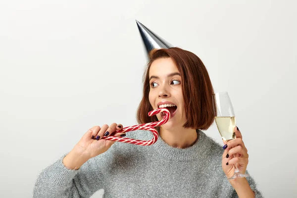 stock image happy woman in party cap holding champagne glass and biting candy cane on grey, Merry Christmas