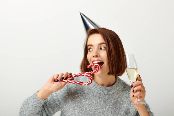 happy woman in party cap holding champagne glass and biting candy cane on grey, Merry Christmas
