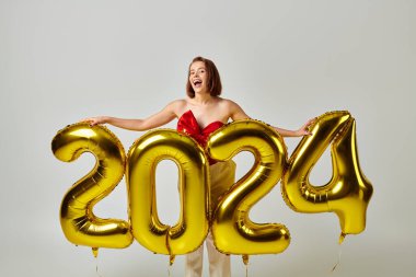 Mutlu yıllar, modaya uygun giyinmiş heyecanlı genç kadın elinde balonlarla gri üzerinde 2024 numara var.
