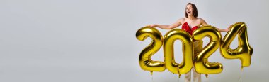 Yeni yıl afişi, modaya uygun giyinmiş heyecanlı genç bir kadın elinde balonlarla gri üzerinde 2024 numara var.