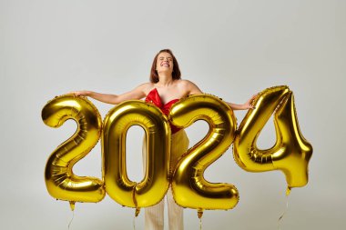 Mutlu yıllar, modaya uygun giyinmiş neşeli genç kadın elinde balonlarla gri üzerinde 2024 numara var.