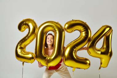 Happy New Year, pleased young woman in trendy attire holding balloons with 2024 numbers on grey clipart