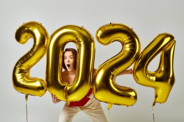 Yeni yılınız kutlu olsun, modaya uygun giyinmiş, elinde 2024 numaralı balon tutan gri elbiseli afallamış genç bayan.