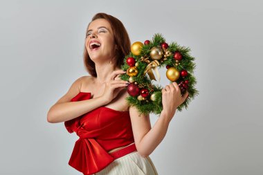 festive season, inspired woman with shimmery makeup holding Christmas wreath on grey backdrop clipart