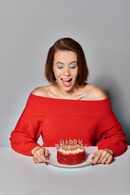 excited woman in red attire looking at bento cake with Happy Birthday candles on grey background clipart