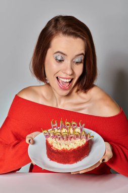 thrilled woman in red attire looking at bento cake with Happy Birthday candles on grey background clipart