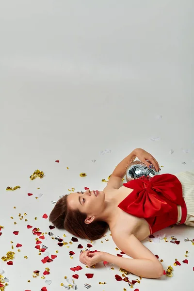 stock image New Year party, brunette woman with shiny disco ball lying on floor near confetti on grey backdrop