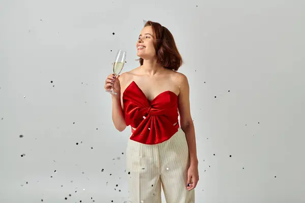 stock image Happy New Year, cheerful woman in party attire holding glass of champagne near confetti on grey