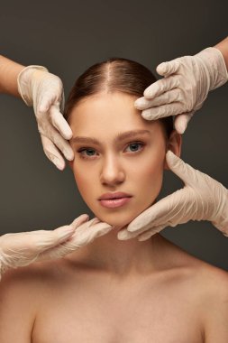 Tıbbi eldivenli estetisyenler gri arka planda genç bir kadının yüzünü inceliyor, dermatoloji konsepti