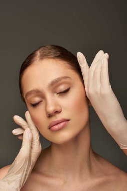 Tıbbi eldivenli estetisyenler gri arka planda kadının yüzüne dokunuyor, dermatoloji konsepti.