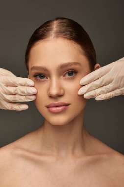 Tıbbi eldivenli estetisyenler gri arka planda güzel bir kadının yüzüne dokunuyor, dermatoloji konsepti.