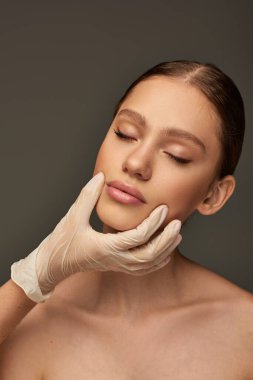 Tıbbi eldivenli estetisyen. Gri arka planda güzel bir kadının yüzüne dokunuyor. Dermatoloji kavramı.