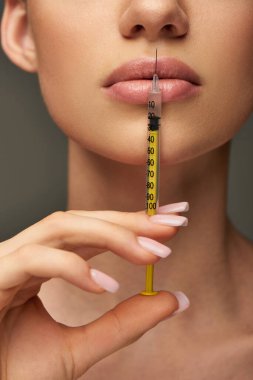 cropped view of young woman holding syringe near face on grey background, lip enhancement concept clipart