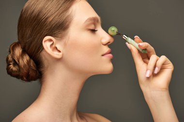 side view of young woman with closed eyes doing massage on nose with jade roller on grey background clipart