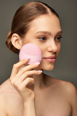 Gri arka planda cilt bakım aleti tutan genç bir kadın, pembe temizlik fırçası.