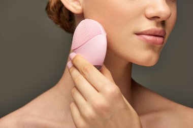 cropped woman holding cleansing brush on grey background, beauty gadget and skin care concept clipart