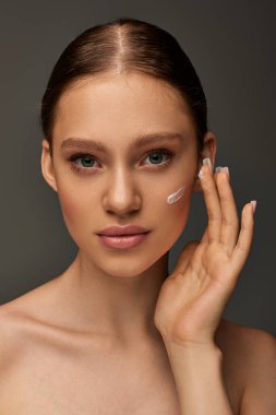 beautiful young woman with bare shoulders applying cream on face on grey background, look at camera clipart