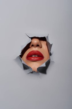 cropped view of young woman with red lips near ripped grey background with hole, peeking clipart