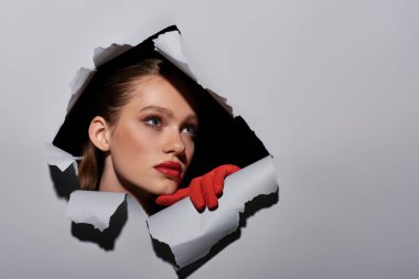 pretty young woman with blue eyes and red lipstick peeking through hole in ripped grey paper clipart