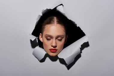 pretty young woman with blue eyes and red lipstick peeking through hole in ripped grey paper clipart
