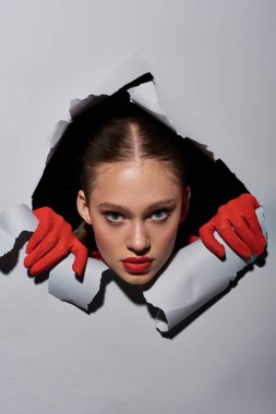 young woman with red lipstick and gloves peeking through hole in ripped grey paper, bold makeup clipart
