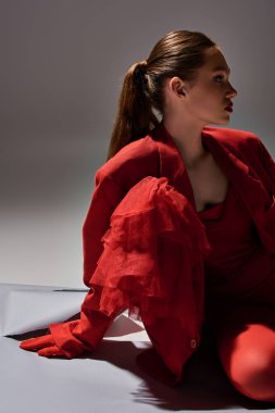 Gri arka planda oturan kırmızı elbiseli çekici genç manken, moda ve tarz sahibi.